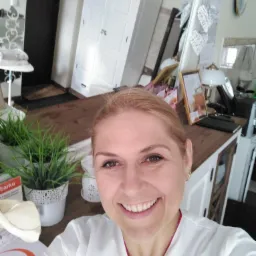 Uśmiechnięta kobieta w białym uniformie, prawdopodobnie pracownik salonu, robi selfie na tle wnętrza z dekoracjami w stylu skandynawskim, widoczne rośliny w doniczkach i lustro.