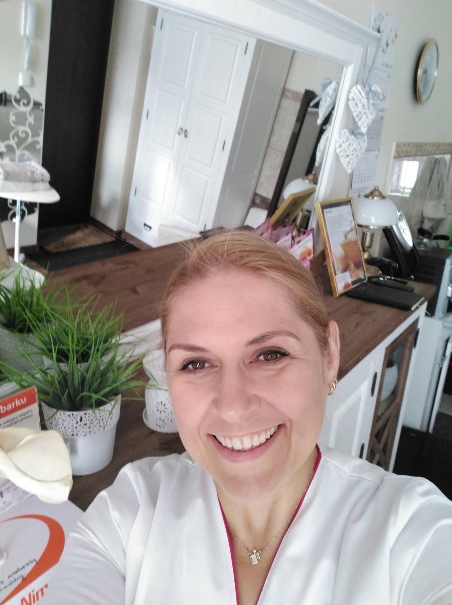 Uśmiechnięta kobieta w białym uniformie, prawdopodobnie pracownik salonu, robi selfie na tle wnętrza z dekoracjami w stylu skandynawskim, widoczne rośliny w doniczkach i lustro.