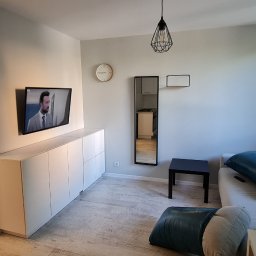 Daad Matter Spółka z Ograniczoną Odpowiedzialnością - Jasne wnętrze salonu z telewizorem na ścianie, białymi szafkami pod nim, lustrem, sofą z poduszkami i lampą wiszącą w stylu industrialnym.