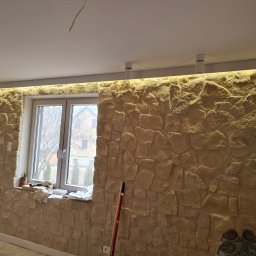 Daad Matter Spółka z Ograniczoną Odpowiedzialnością - Wykończona ściana z kamienia dekoracyjnego wewnątrz pomieszczenia, z oknem i ukrytym oświetleniem LED w suficie podwieszanym, widoczne narzędzia i akcesoria na parapecie.