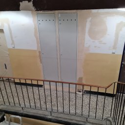 Daad Matter Spółka z Ograniczoną Odpowiedzialnością - Klatka schodowa w bloku, ściany w trakcie remontu, widoczne dwie szare skrzynki elektryczne z otworami wentylacyjnymi, zabezpieczone drzwi do mieszkań.