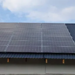 Panele fotowoltaiczne zamontowane na dachu z blachy trapezowej, widoczne jasne chmury na błękitnym niebie w tle.