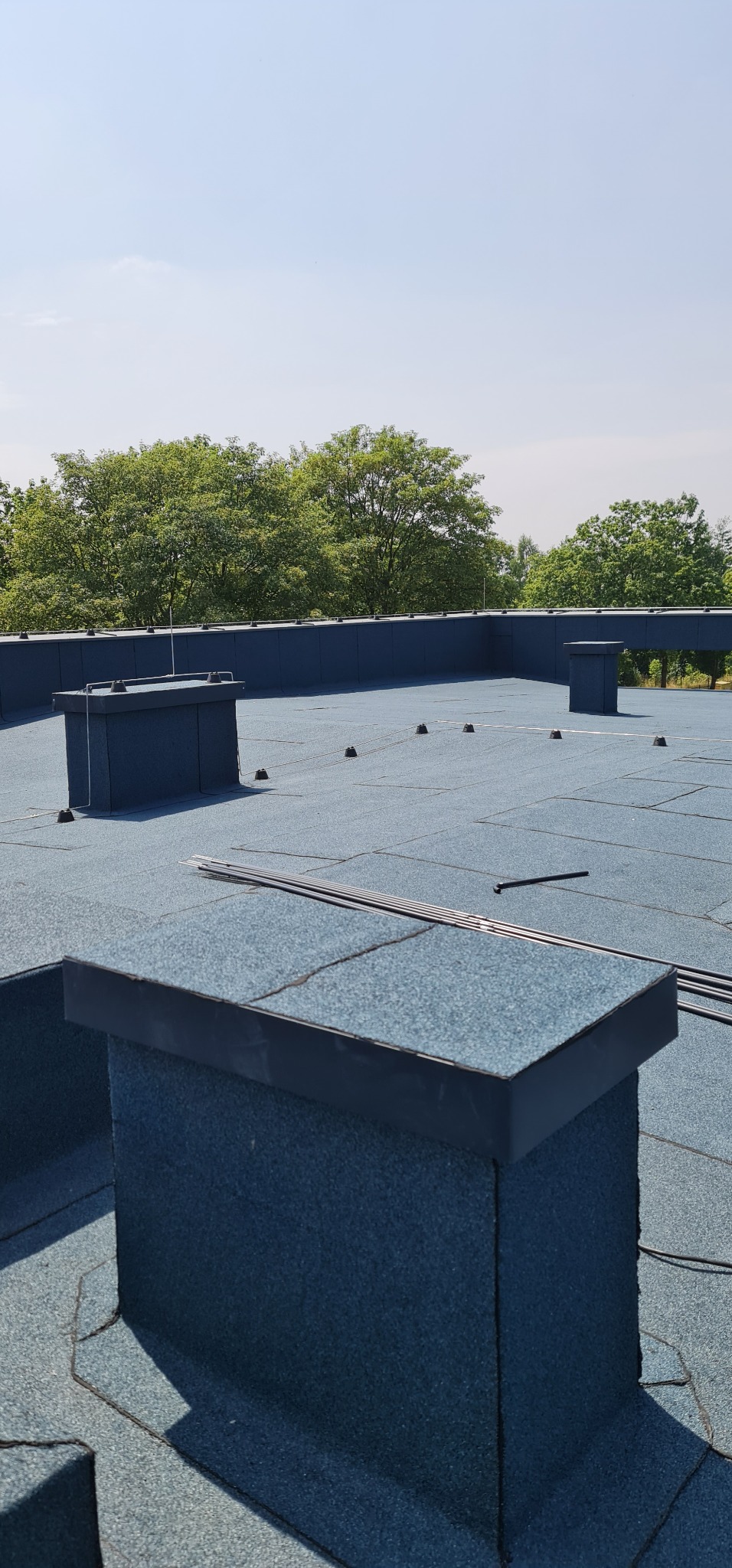 Instalacja odgromowa na płaskim dachu pokrytym papą, widoczne elementy systemu uziemiającego i przewody odprowadzające, tło stanowią drzewa i błękitne niebo.