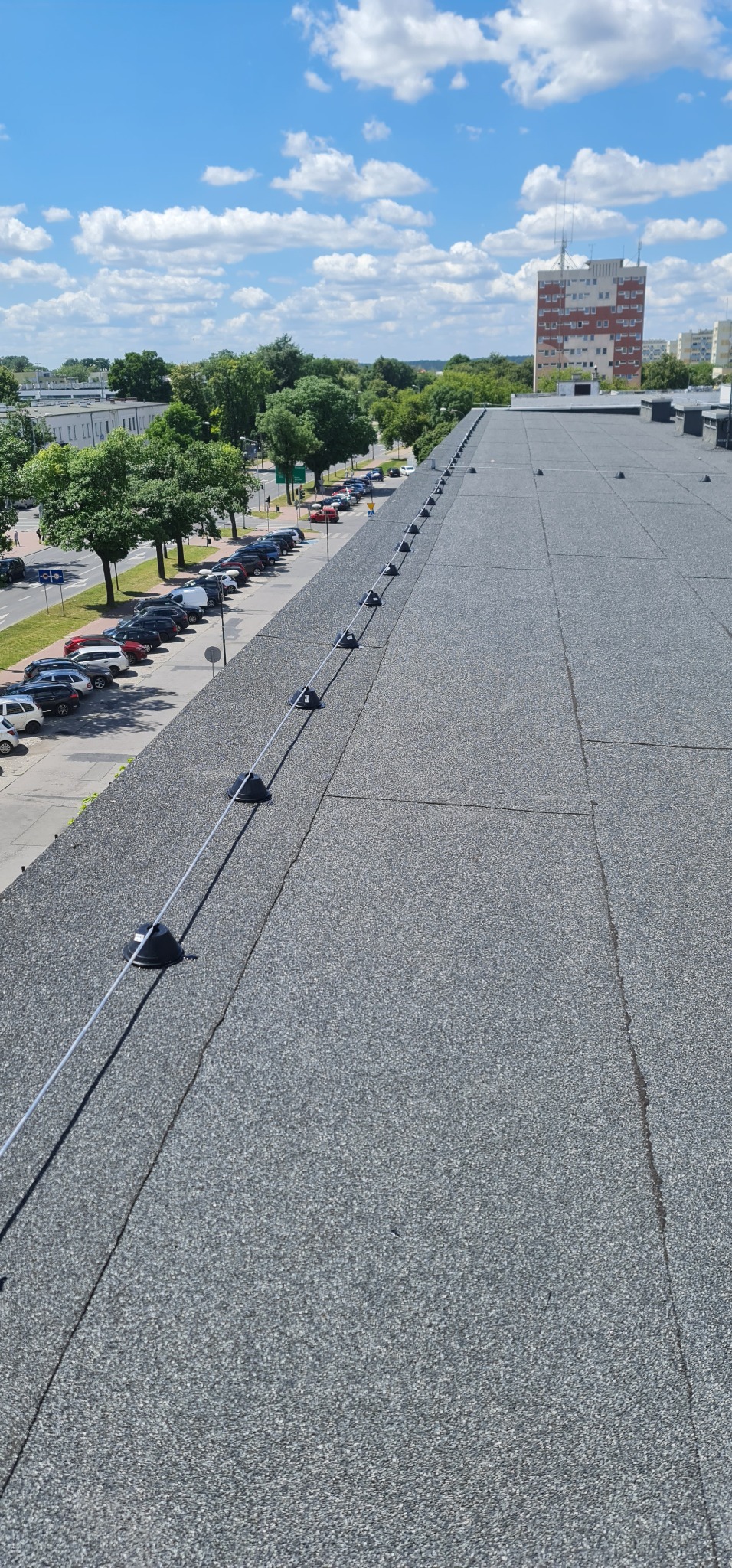 Widok z góry na płaski dach pokryty papą z zamontowaną instalacją odgromową, na tle zabudowań miejskich i błękitnego nieba z chmurami.