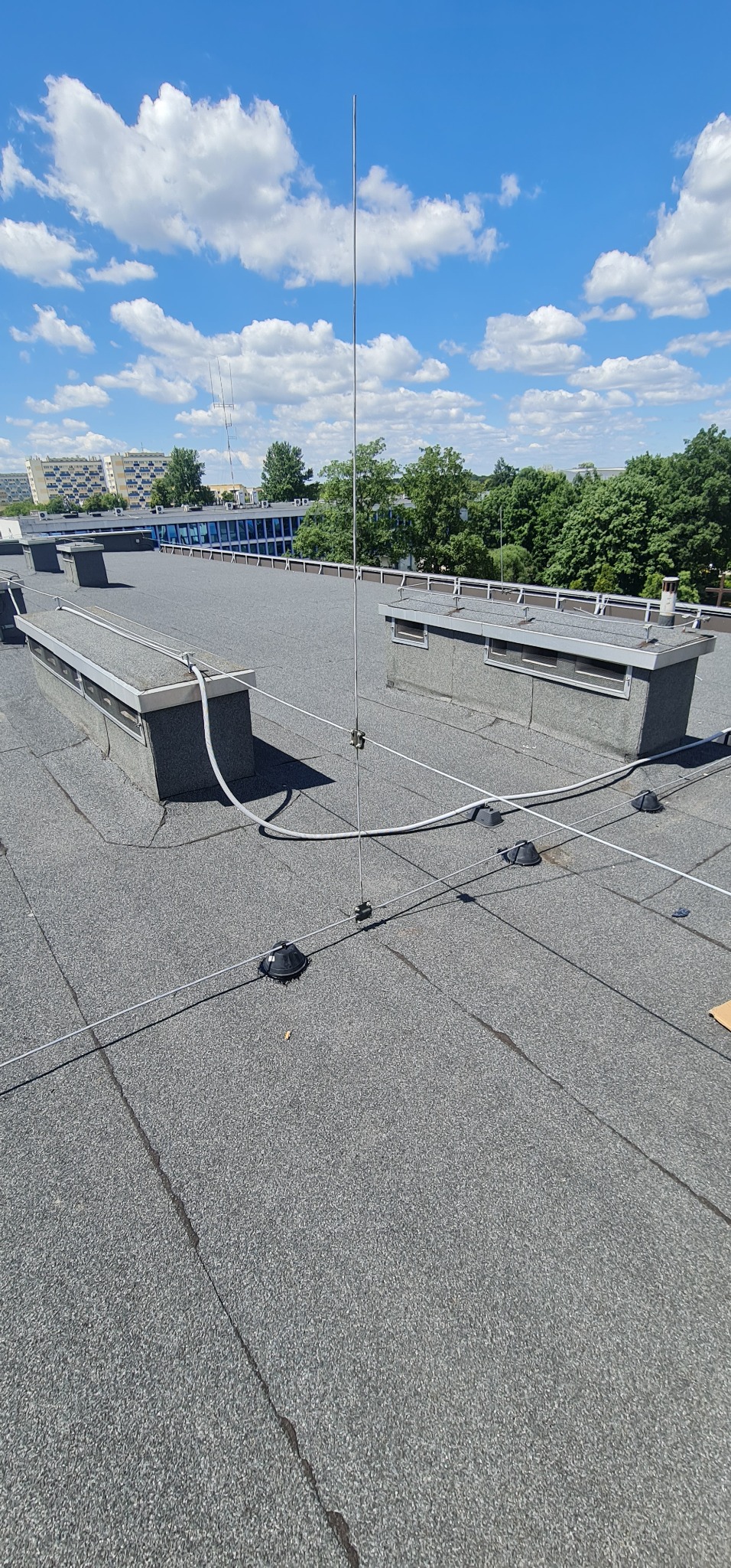 System ochrony odgromowej z pionowym masztem i przewodami uziemiającymi zamontowany na płaskim dachu budynku z widocznymi elementami mocującymi i tłem błękitnego nieba.