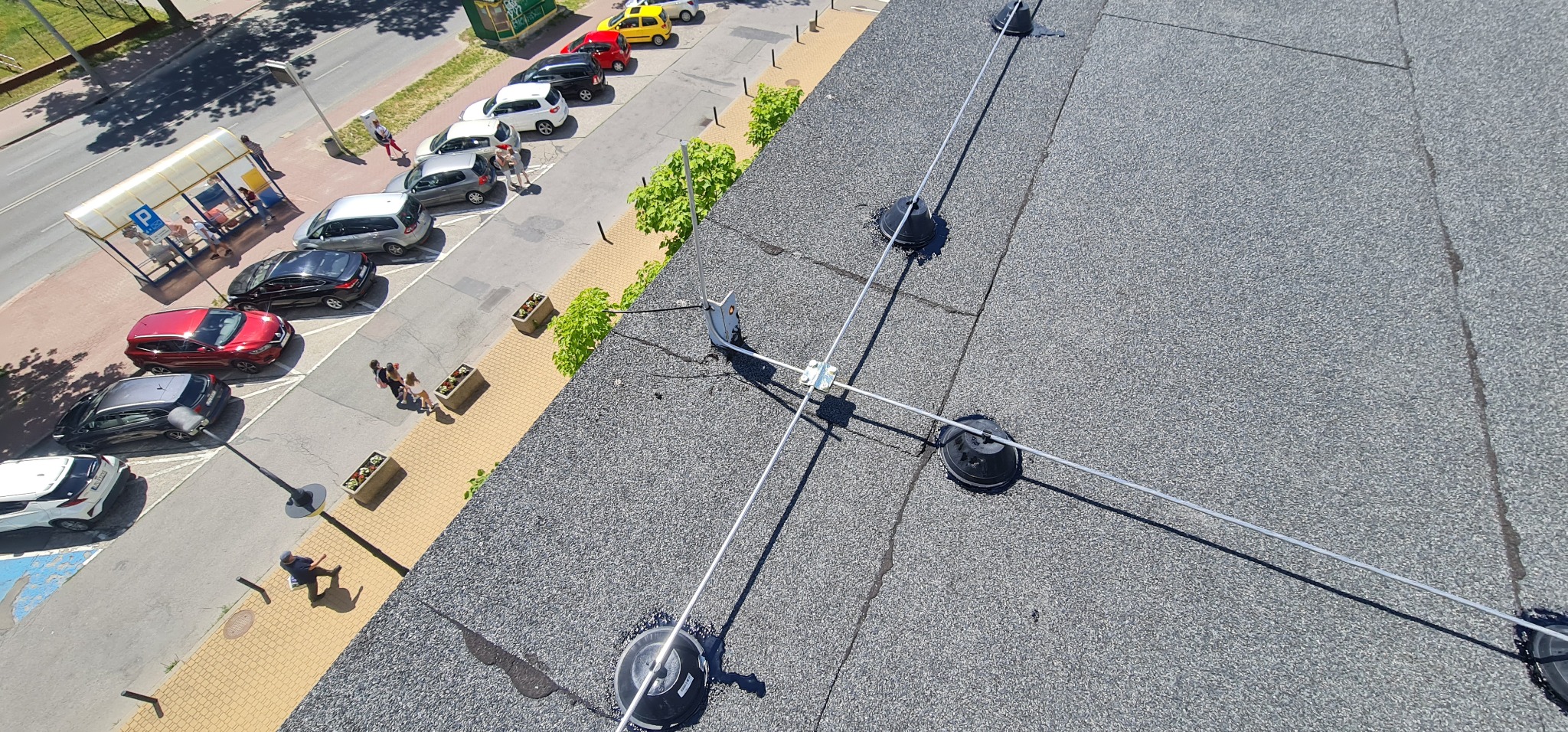 Widok z góry na fragment dachu pokrytego papą z zainstalowaną instalacją odgromową z trzema uchwytami i przewodami, na tle chodnika, parkingu i ulicy z przechodniami i samochodami w słoneczny dzień.