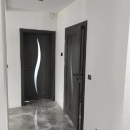 Dwa ciemnoszare drzwi wewnętrzne z szybą o falistym kształcie w nowoczesnym korytarzu z szarą podłogą z płytek imitujących marmur.