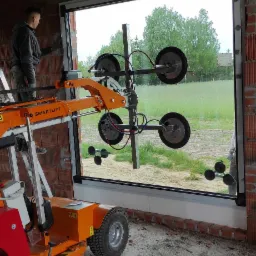 Montaż okna przy użyciu podnośnika próżniowego Smartlift w surowym wnętrzu budynku z widokiem na zewnątrz.