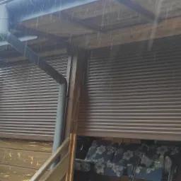 Zasłonięte brązowe rolety zewnętrzne w drewnianej altanie podczas deszczu, widoczne rynny i fragment sąsiedniego domu.