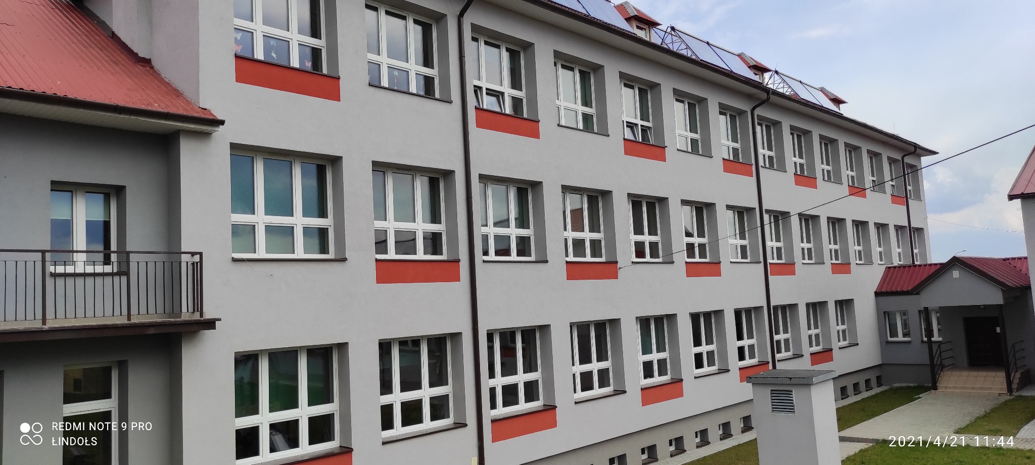 Szary budynek szkoły z białymi oknami, czerwonymi akcentami i panelami słonecznymi na dachu, sfotografowany w pochmurny dzień.