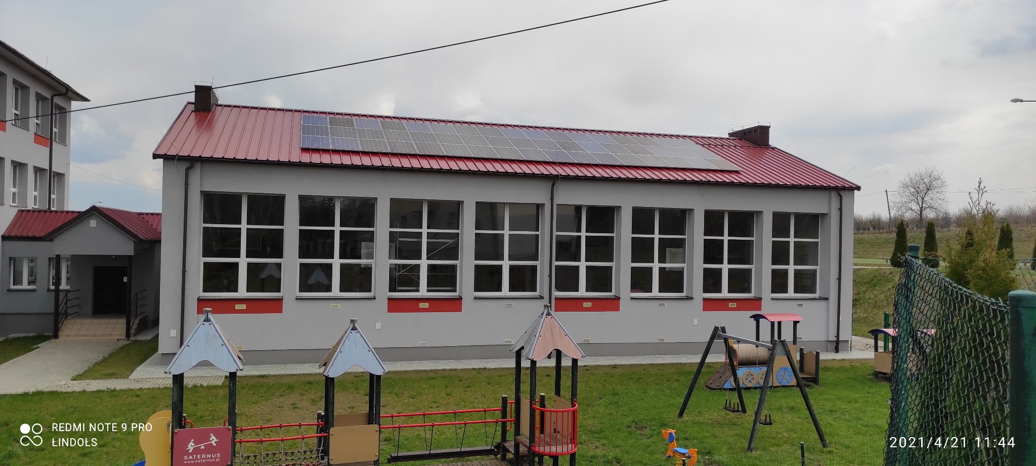 Szary budynek szkoły z czerwonym dachem i panelami słonecznymi, widok z placu zabaw z elementami wyposażenia.