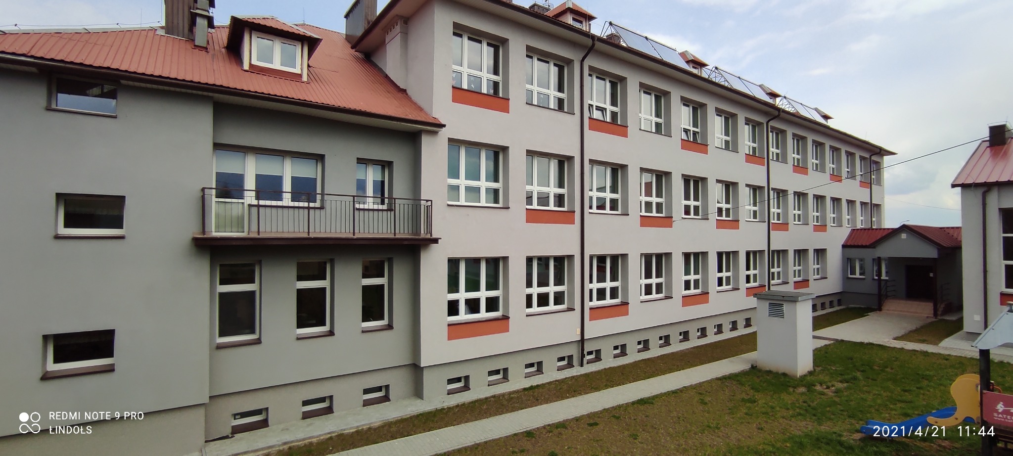 Szary budynek z białymi oknami PCV i panelami fotowoltaicznymi na dachu, widok z zewnątrz w pochmurny dzień.