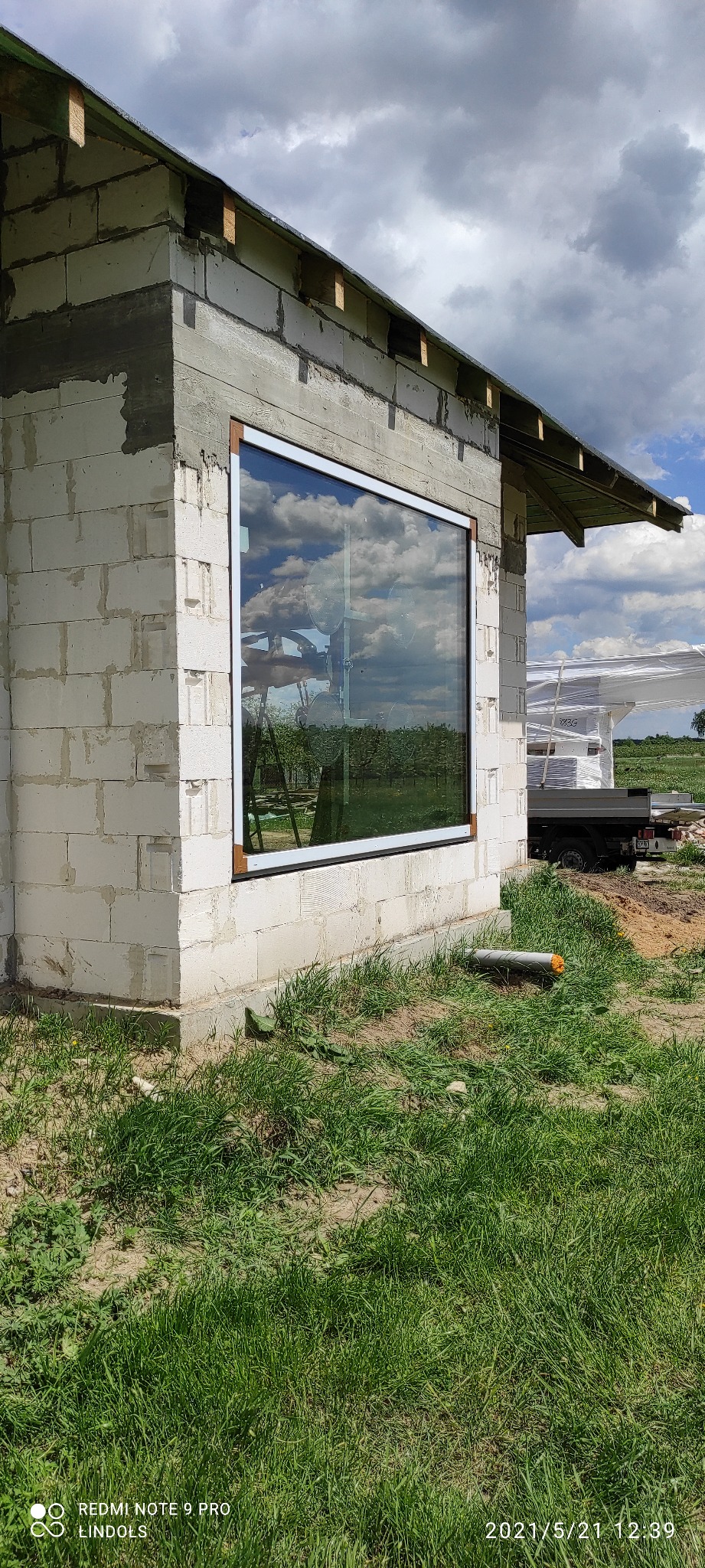 Montaż dużego okna z białą ramą w surowej ścianie z bloczków, odbicie chmur i drzew w szybie, widoczny podnośnik do szyb i przyczepa z materiałami budowlanymi w tle.