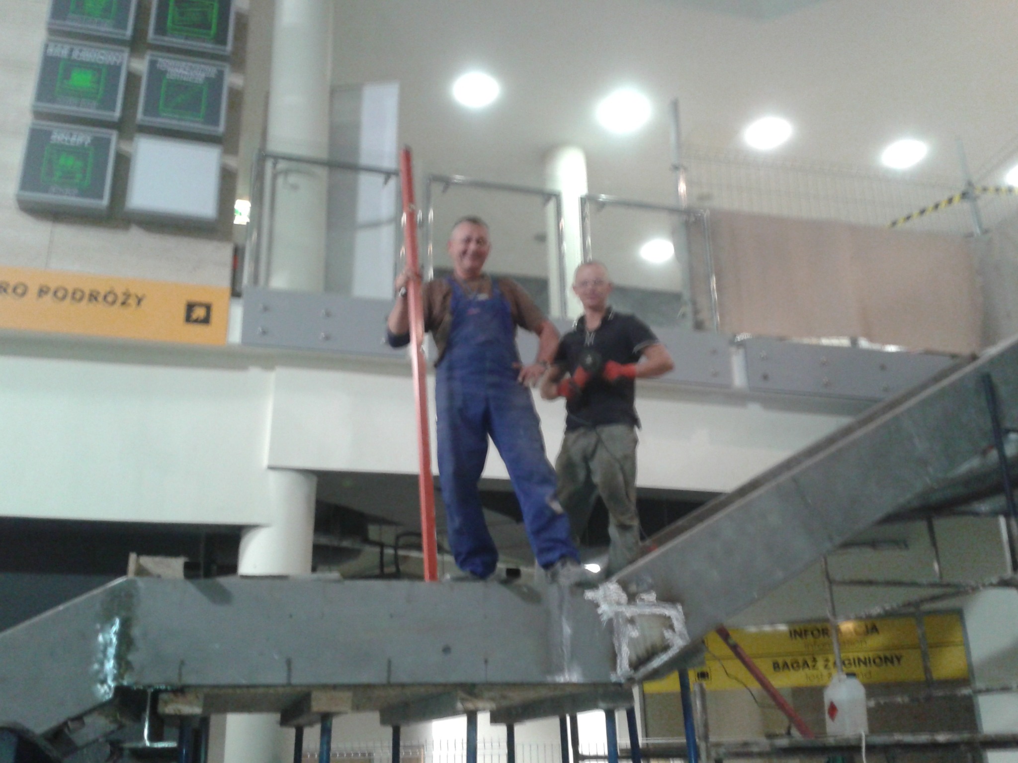 Dwóch mężczyzn w roboczych ubraniach na metalowej konstrukcji w hali, jeden trzyma poziomicę, drugi z narzędziem w ręku, w tle tablice informacyjne i rusztowania.