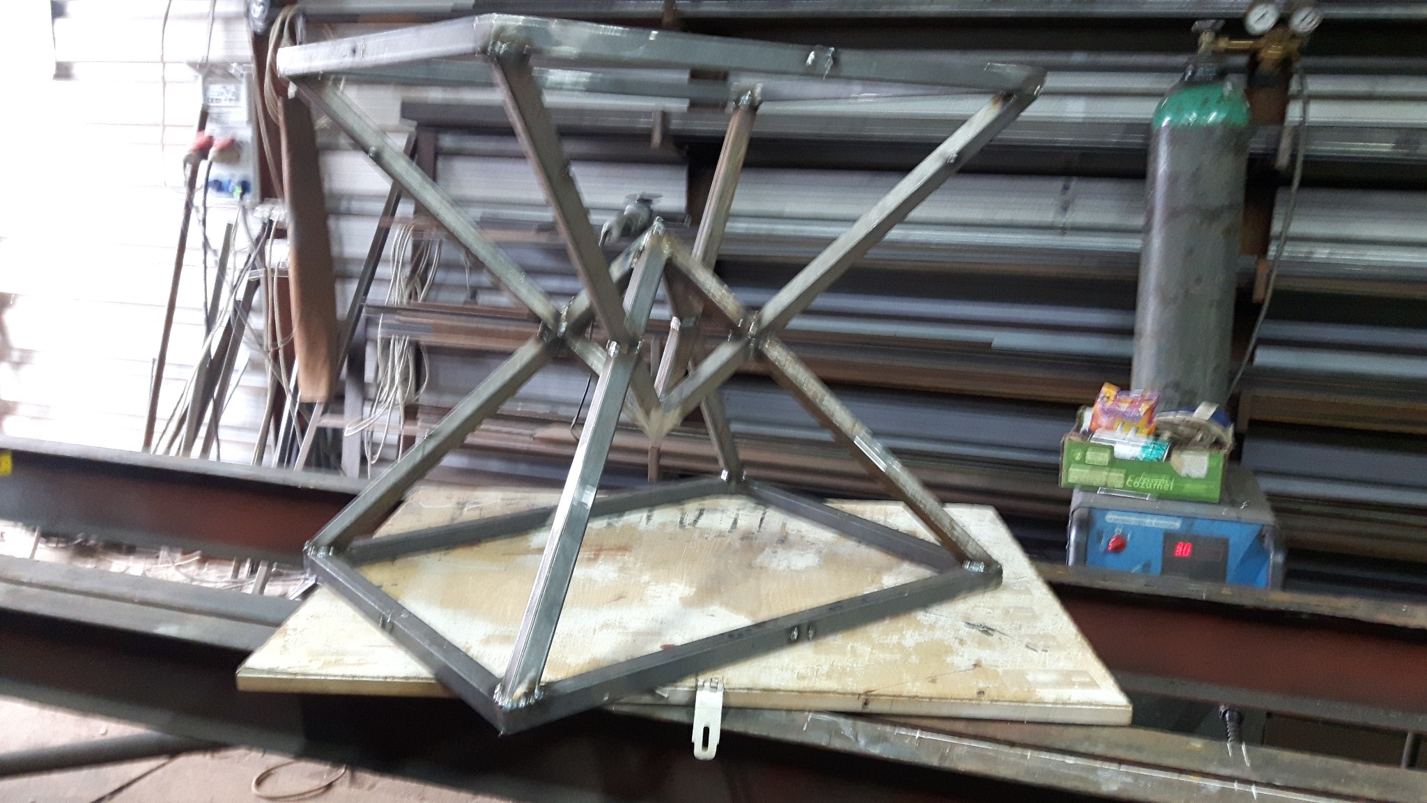 Konstrukcja metalowa w kształcie klepsydry z kwadratowymi podstawami, spawana z profili stalowych, umieszczona na drewnianej płycie w warsztacie, widoczny butla z gazem i spawarka w tle.