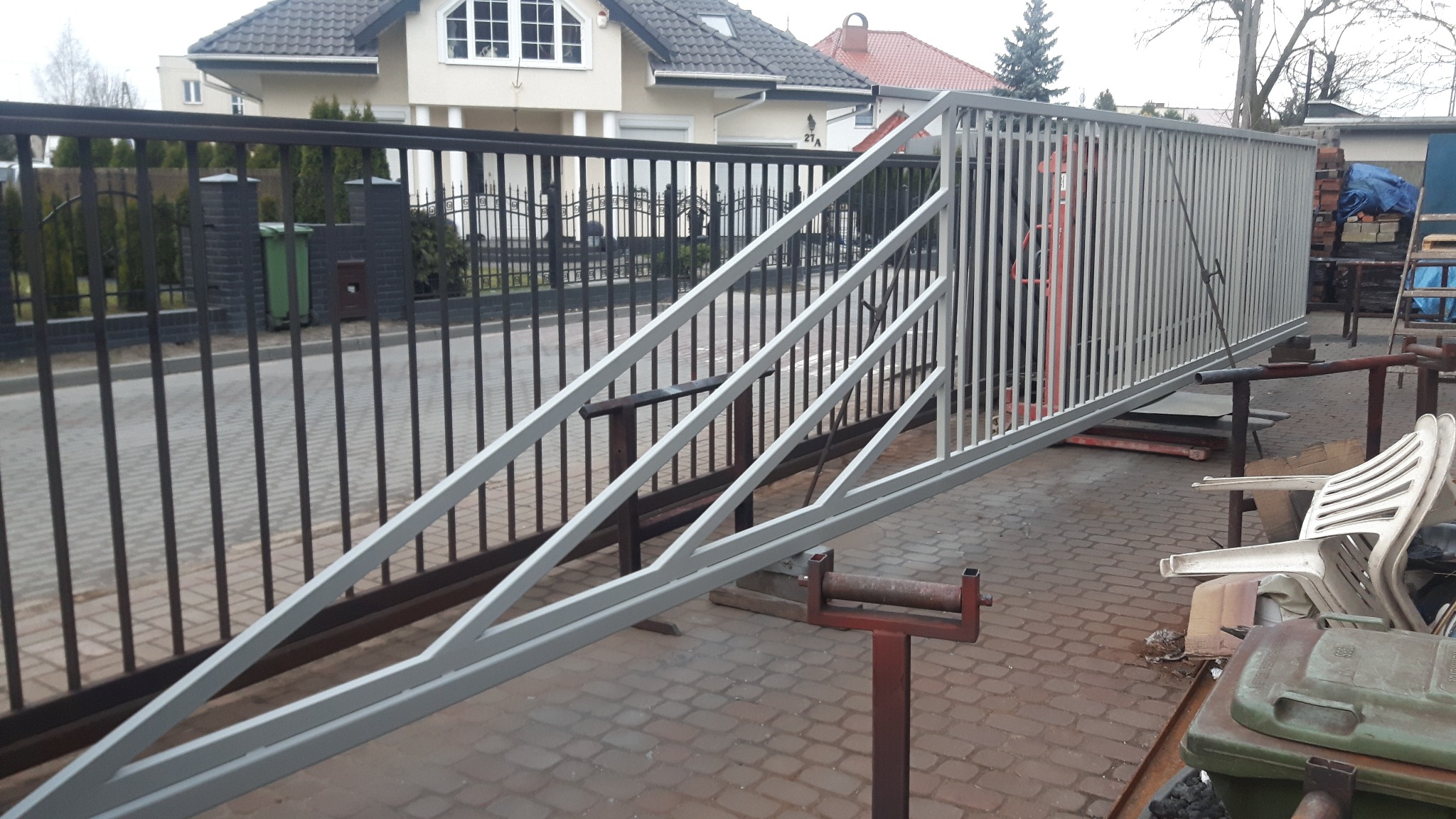 Fragment metalowej konstrukcji ogrodzenia panelowego w warsztacie, widoczny spaw na łączeniu elementów, w tle czarne ogrodzenie posesji i budynki mieszkalne.