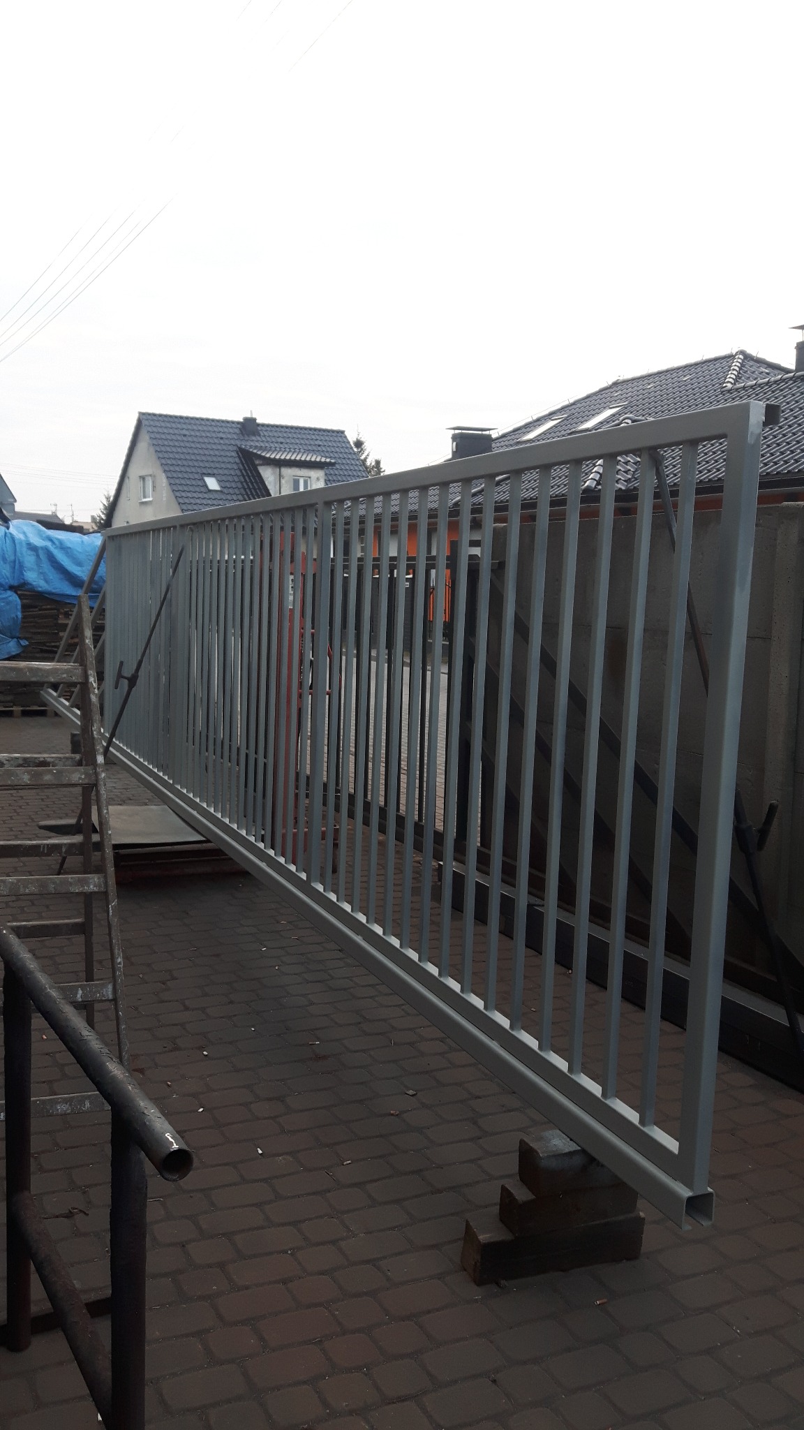 Szary panel ogrodzeniowy oparty na drewnianych klockach, widoczna drabina i kostka brukowa na tle zabudowań.