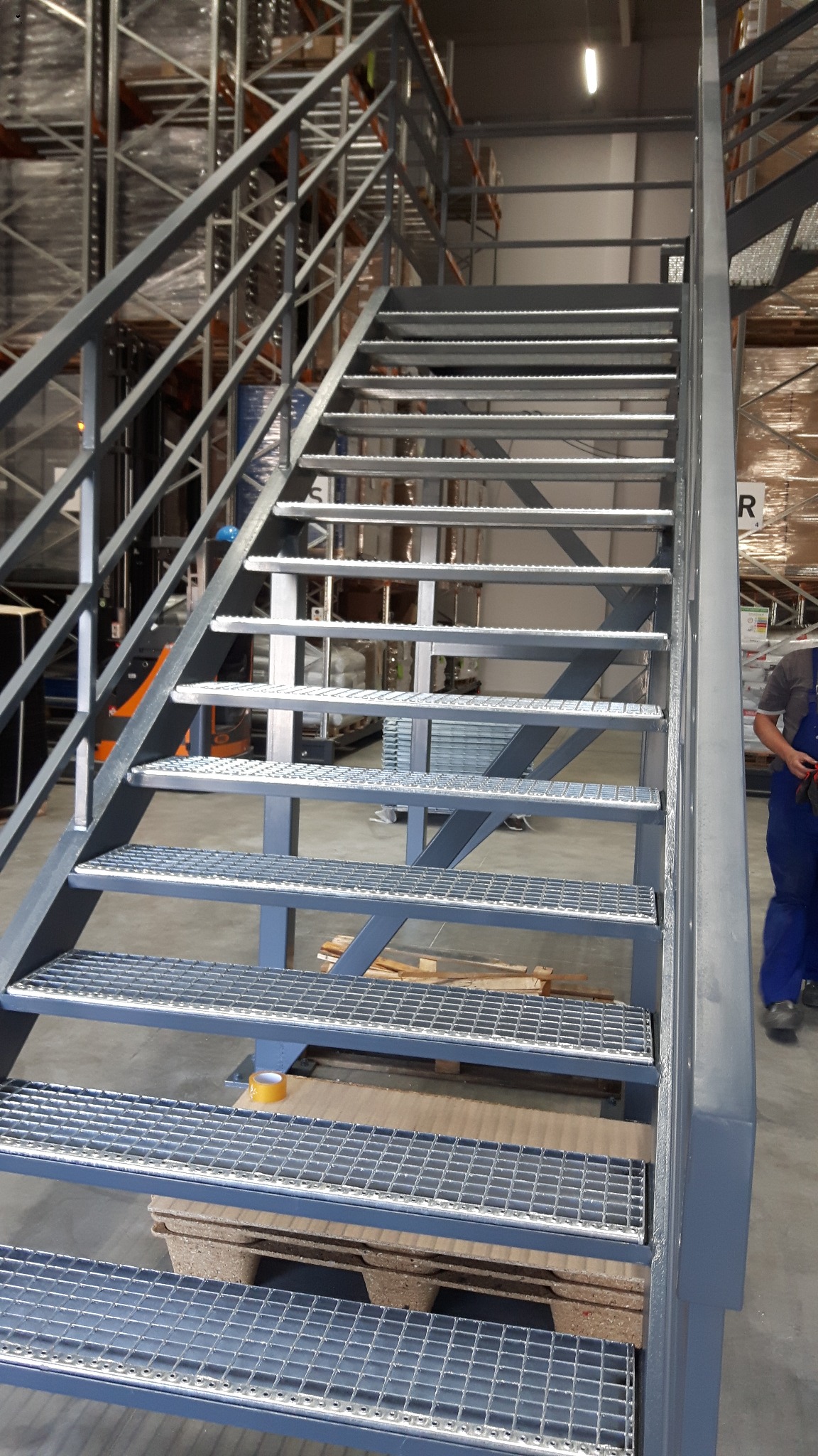 Metalowe schody kratowe w magazynie, widoczne regały w tle, taśma klejąca na jednym ze stopni.