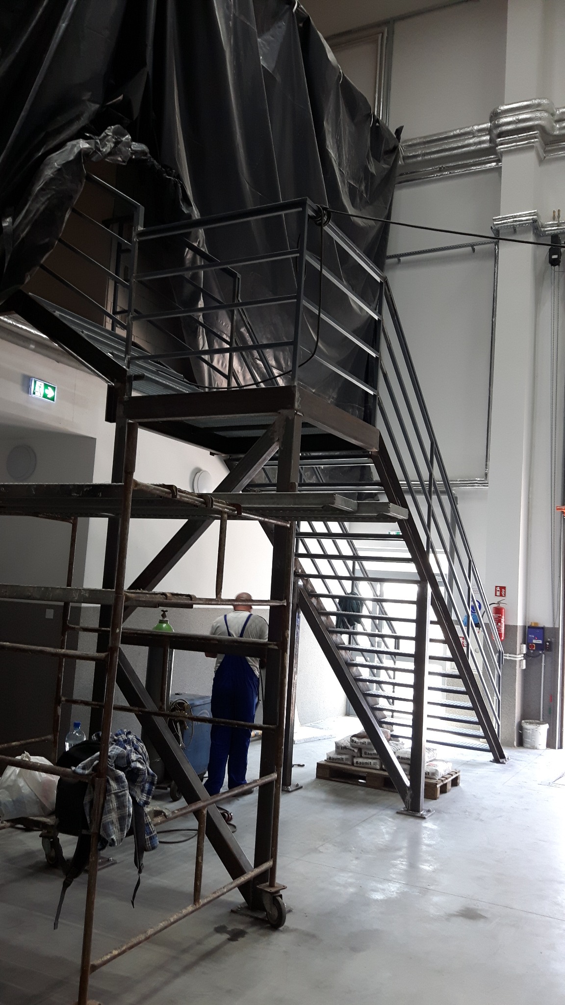 Montaż metalowej konstrukcji schodów z platformą, widoczne rusztowanie, pracownik w niebieskim kombinezonie oraz materiały budowlane na palecie.