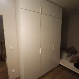 Wysoka, biała szafa z prostymi uchwytami, sięgająca sufitu w sypialni, widoczna przy ścianie obok łóżka.