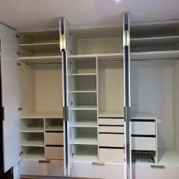 Wnętrze trzydrzwiowej, białej szafy wnękowej z półkami, drążkami na wieszaki i szufladami, widoczne otwarte drzwi i metalowe uchwyty.