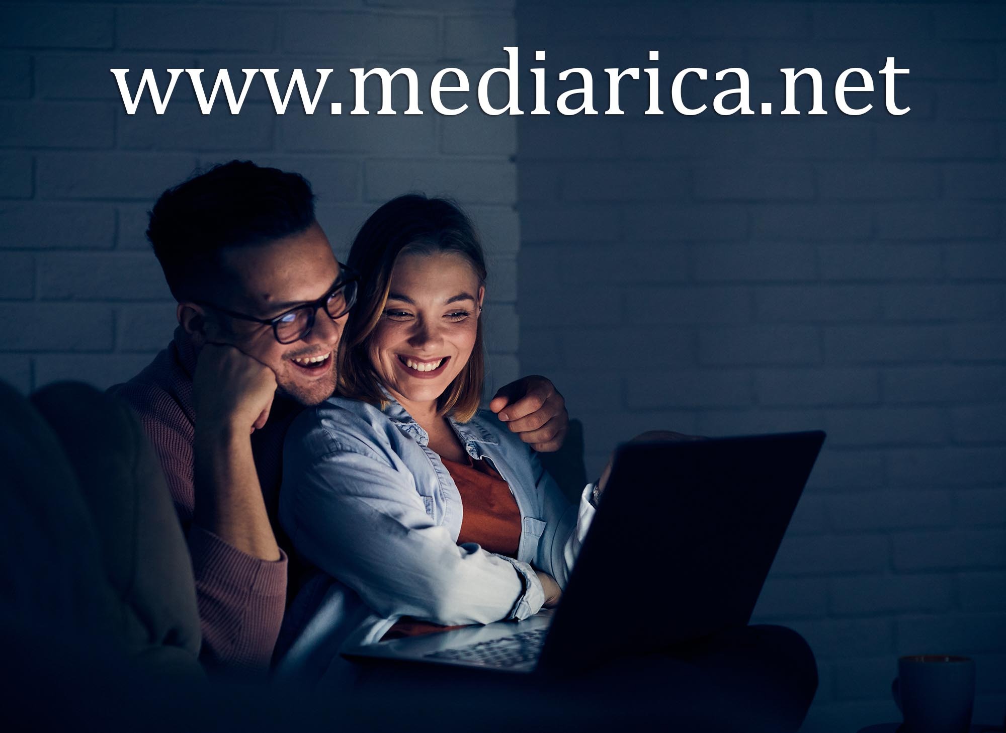 Uśmiechnięta para w okularach ogląda coś na laptopie w słabo oświetlonym pokoju z ceglaną ścianą w tle, na ekranie laptopa widoczny blask.