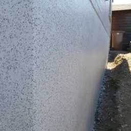 Ocieplona ściana budynku z wykończeniem strukturalnym w odcieniach bieli i szarości, widoczna z boku, z fragmentem okna i rynny oraz ziemią i drobnymi kamieniami u podstawy.