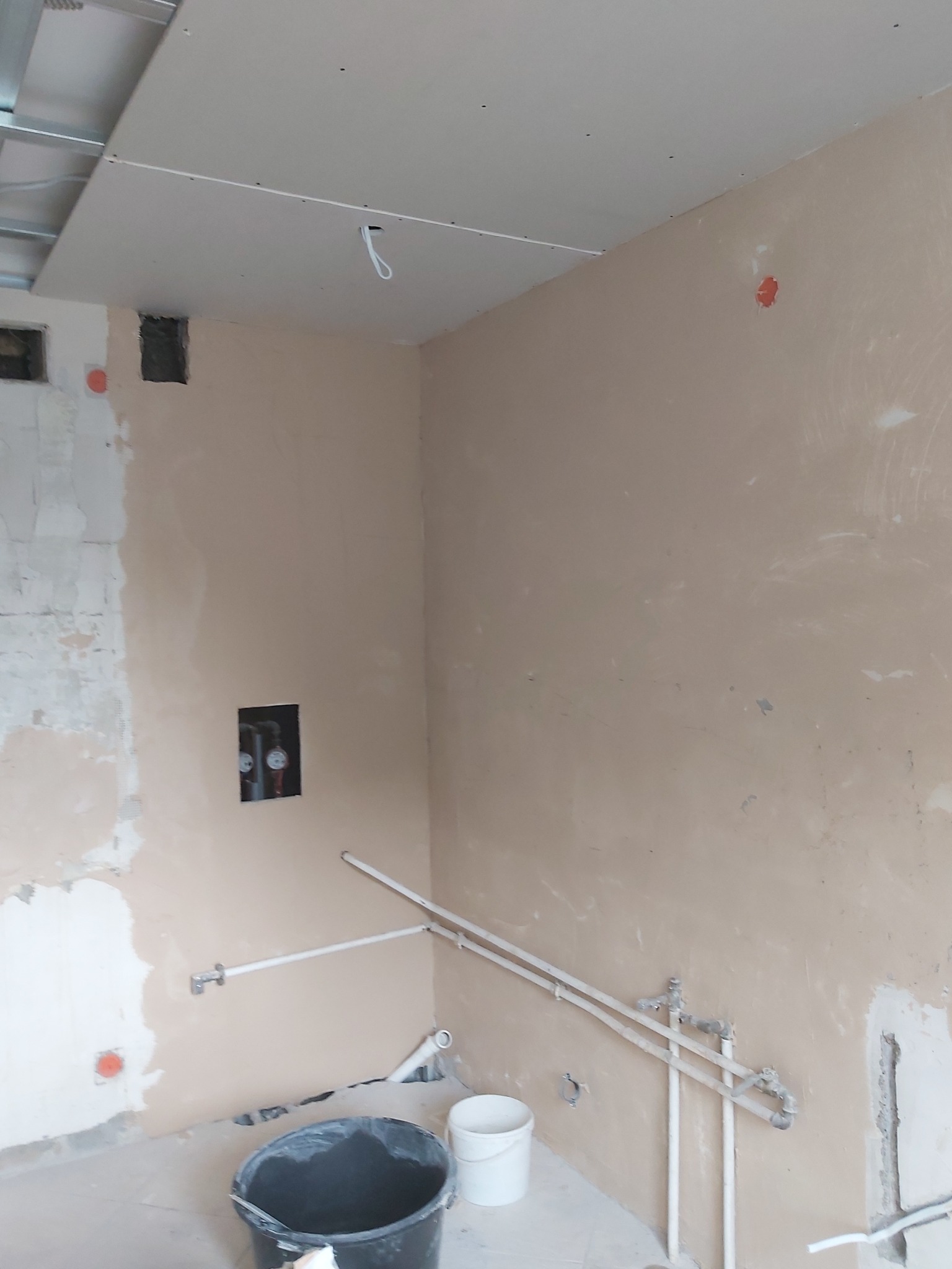 Wnętrze w trakcie prac wykończeniowych: ściany pokryte gładzią, widoczne rury instalacji wodnej, otwory pod gniazdka elektryczne, sufit podwieszany w trakcie montażu, wiadra budowlane na podłodze.