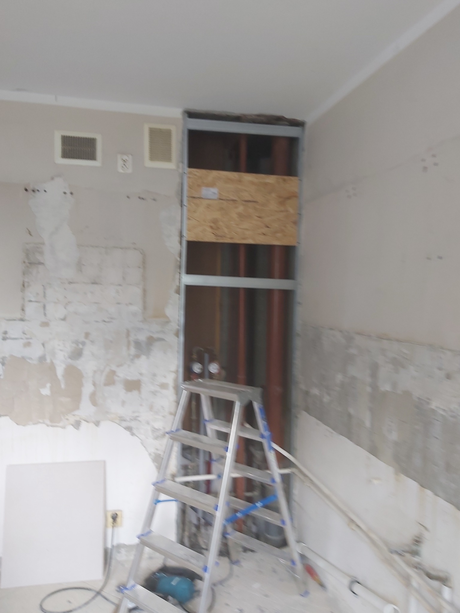 Wnętrze w trakcie prac remontowych, ściany z widocznymi śladami skuwania, odsłonięte rury w metalowej ramie, drabina aluminiowa i płyta gipsowa w tle.
