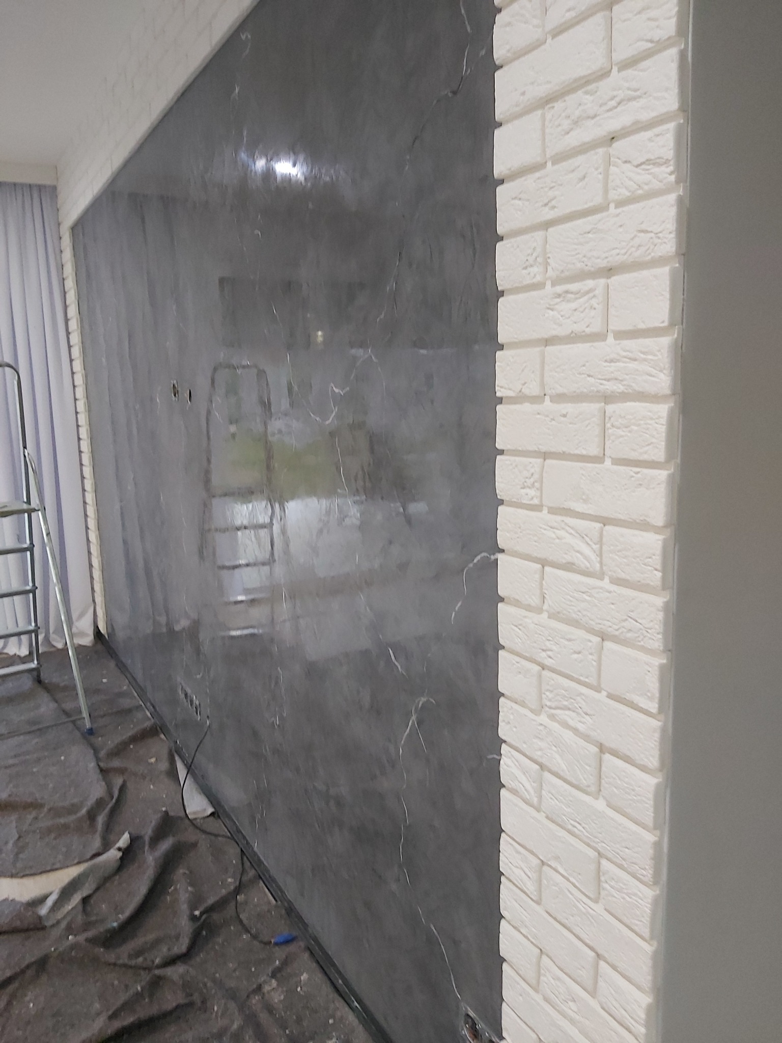 Ściana z efektem marmuru w odcieniach szarości sąsiadująca z fragmentem ściany wyłożonej dekoracyjną białą cegłą, widoczna drabina malarska i zabezpieczona podłoga.