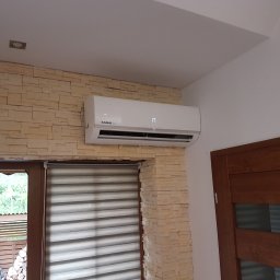 T-WENT Piotr Tenderenda - Biały klimatyzator Kaisai zamontowany na ścianie z dekoracyjnej cegły, widoczny fragment okna z żaluzjami i drzwi.