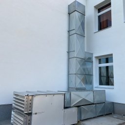 T-WENT Piotr Tenderenda - Zewnętrzny montaż systemu rekuperacji z pionowym kominem wentylacyjnym na tle białej ściany budynku z oknami, widoczne żółte kable.