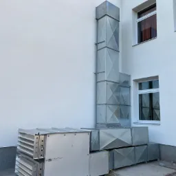 Zewnętrzny montaż systemu rekuperacji z pionowym kominem wentylacyjnym na tle białej ściany budynku z oknami, widoczne żółte kable.