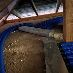 T-WENT Piotr Tenderenda - Niebieskie, karbowane rury wentylacyjne ułożone wzdłuż poddasza, z widoczną drewnianą konstrukcją dachu i fragmentami metalowych kanałów.