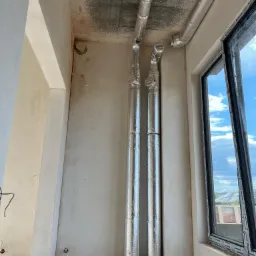 Dwie pionowe rury wentylacyjne w izolacji aluminiowej, prowadzone wzdłuż ściany w surowym stanie, z widocznym fragmentem instalacji pod sufitem i oknem z widokiem na niebo.