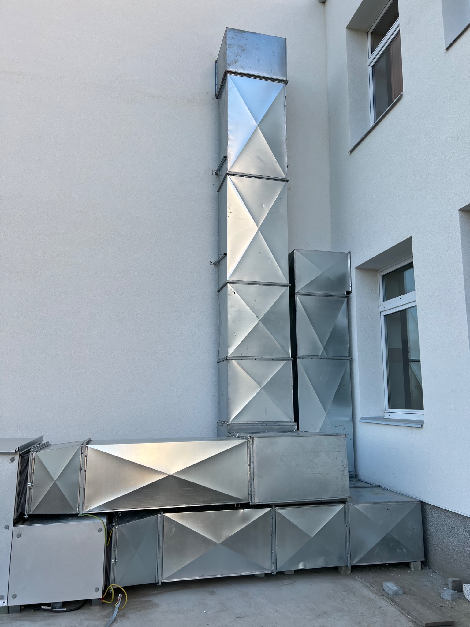 Metalowe kanały wentylacyjne ułożone na ziemi i zamontowane na elewacji budynku, widoczne okna, elementy instalacji elektrycznej.