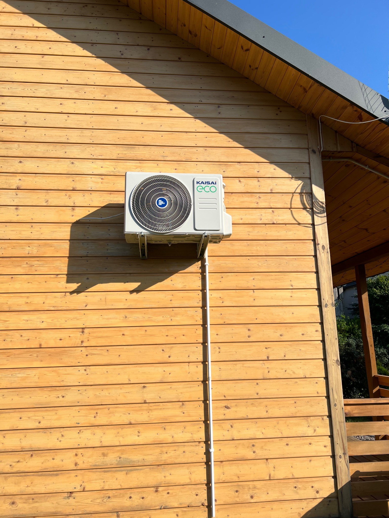 Biała jednostka zewnętrzna klimatyzatora marki Kaisai zamontowana na drewnianej elewacji budynku, z widocznymi przewodami w białej osłonie.
