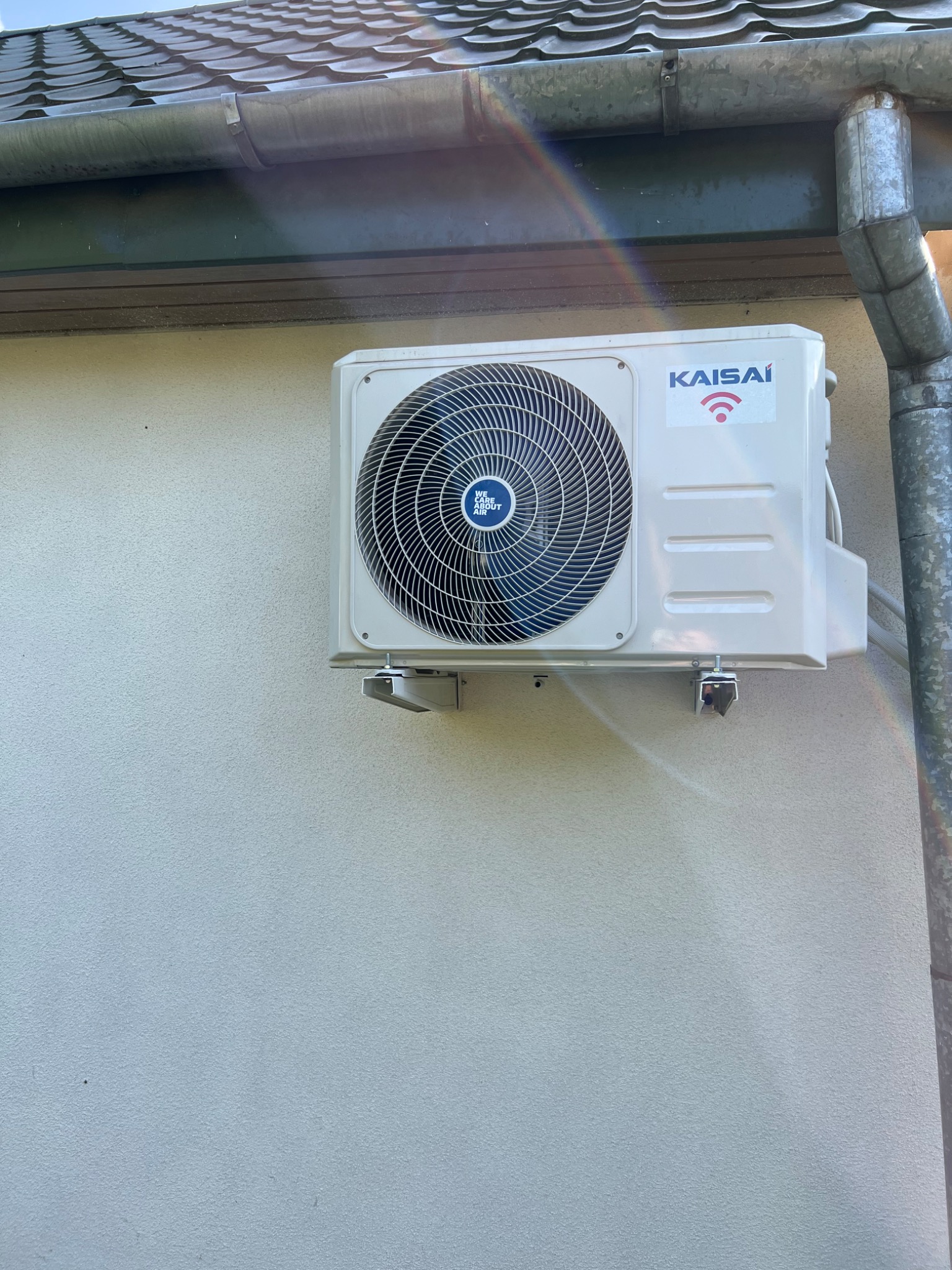 Zewnętrzna jednostka klimatyzacji Kaisai zamontowana na jasnej, teksturowanej ścianie budynku, widoczny fragment rynny i rury spustowej, delikatna tęcza na ścianie.