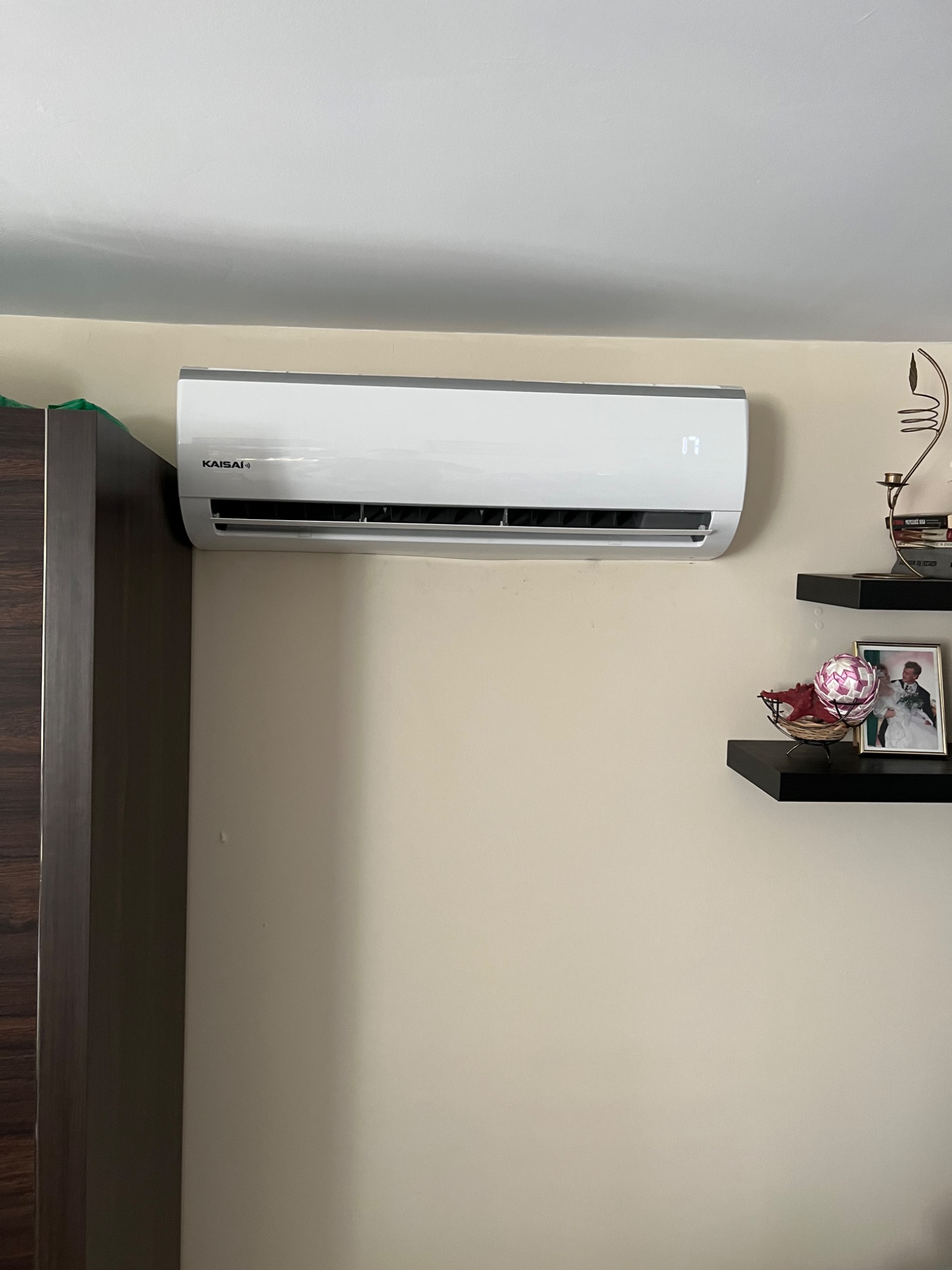 Biały klimatyzator marki Kaisai zamontowany na beżowej ścianie w pomieszczeniu, wyświetlający temperaturę 17 stopni, obok ciemne półki z dekoracjami.