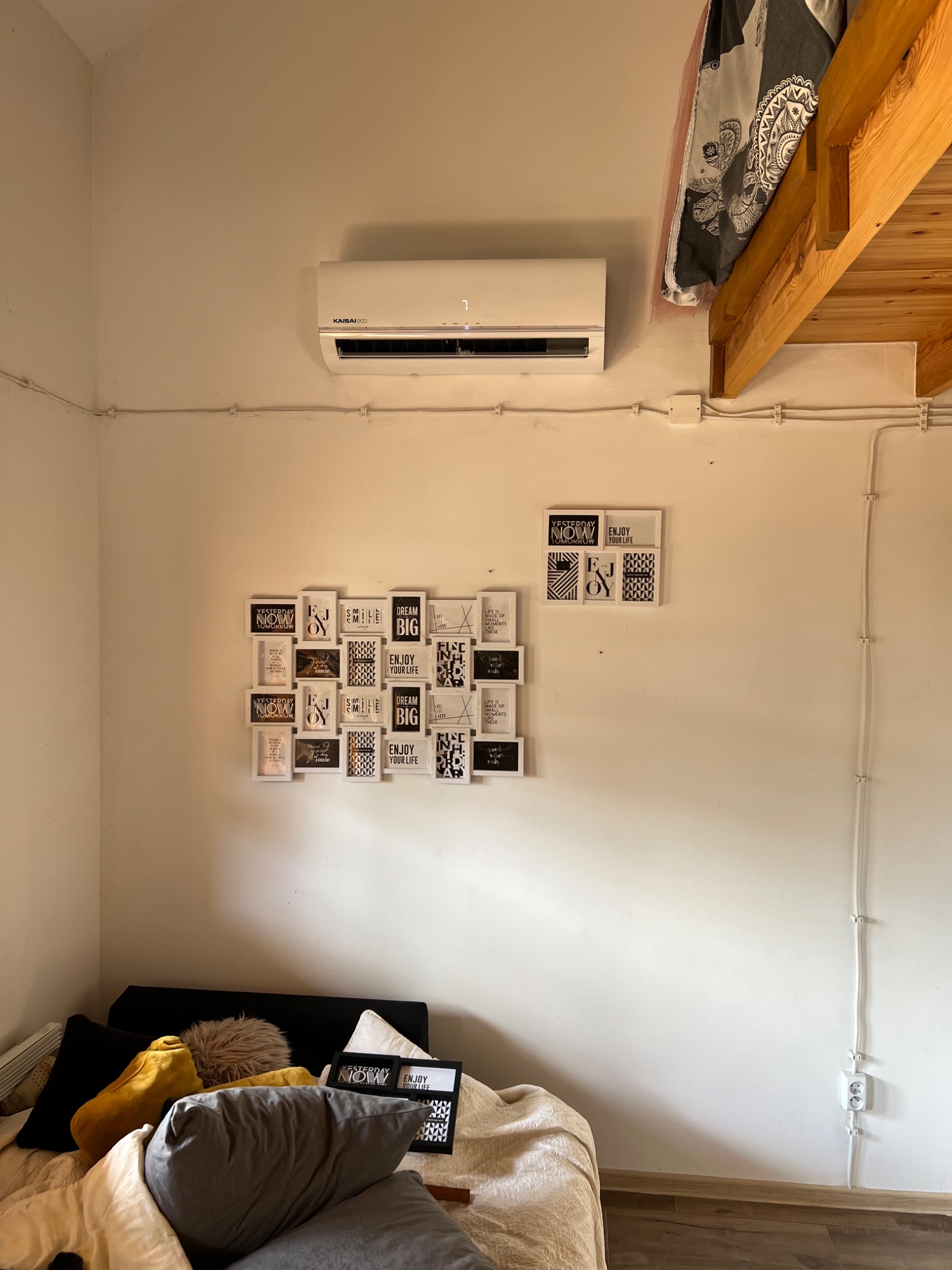 Wnętrze pokoju z zamontowaną klimatyzacją Kaisai Eco na ścianie, pod spodem dekoracja z ramek z napisami motywacyjnymi, widoczne kable elektryczne przytwierdzone do ściany i fragment łóżka...