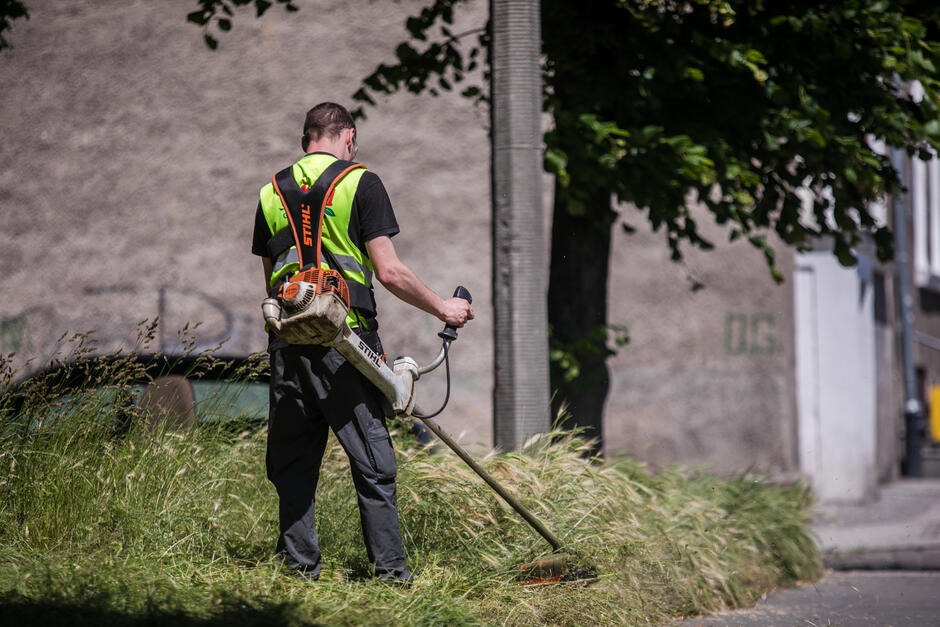 Mężczyzna w kamizelce odblaskowej kosi trawę przy chodniku za pomocą podkaszarki spalinowej Stihl, na tle szarego budynku z zielonym napisem.