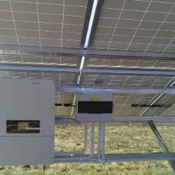 Widok od spodu na panele słoneczne zamontowane na metalowej konstrukcji, z widocznym inwerterem Sofar i skrzynką przyłączeniową, na tle zielonej trawy.