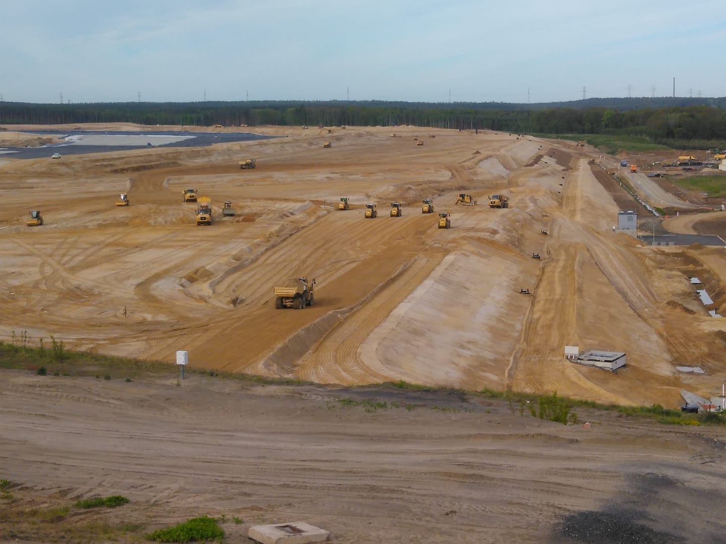 Rozległy teren budowy z licznymi maszynami ziemnymi, w tym wywrotkami i spychaczami, pracującymi na piaszczystym podłożu, z lasem w tle.