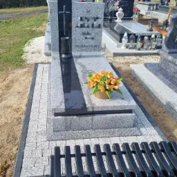 Granitowy pomnik z czarnym pasem i krzyżem, otoczony jasnoszarą kostką brukową i czarną ławką, z bukietem pomarańczowych róż w doniczce.