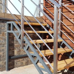 SPEED balustradydlaciebie.pl - Zewnętrzne schody z drewnianymi stopniami i metalową konstrukcją nośną oraz poręczami z rurek, na tle elewacji z kamienia i drewnianego płotu.