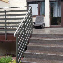 SPEED balustradydlaciebie.pl - Zewnętrzne schody z szarymi płytkami prowadzące na taras z poziomą, szarą balustradą, na tarasie krzesło ogrodowe, w tle okna i drzwi balkonowe.