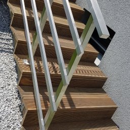 SPEED balustradydlaciebie.pl - Drewniane schody zewnętrzne z nowoczesną, minimalistyczną balustradą z profili o przekroju kwadratowym, zamontowaną do ściany budynku wykończonego szarym tynkiem strukturalnym, u podstawy wysypane...