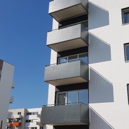 SPEED balustradydlaciebie.pl - Nowoczesny budynek wielorodzinny z metalowymi balkonami z perforowanymi ściankami, widok z dołu na tle błękitnego nieba, plac budowy w trakcie wykańczania.