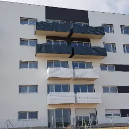 Nowoczesny budynek mieszkalny z balkonami o perforowanych, ciemnoszarych i białych panelach, widok z dołu na tle błękitnego nieba.