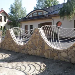 Falista, niska konstrukcja ogrodzeniowa z kamienną podstawą i białymi, giętymi metalowymi prętami w kształcie fali, otaczająca posesję z domami w tle.